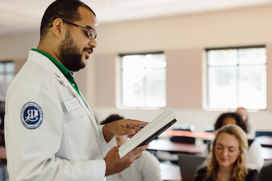 a male student standing in a classroom reading from a textbook